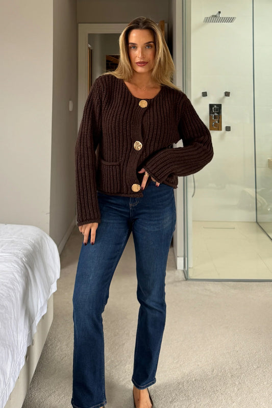 Woman wearing a brown ribbed sweater and blue jeans in a bedroom setting.