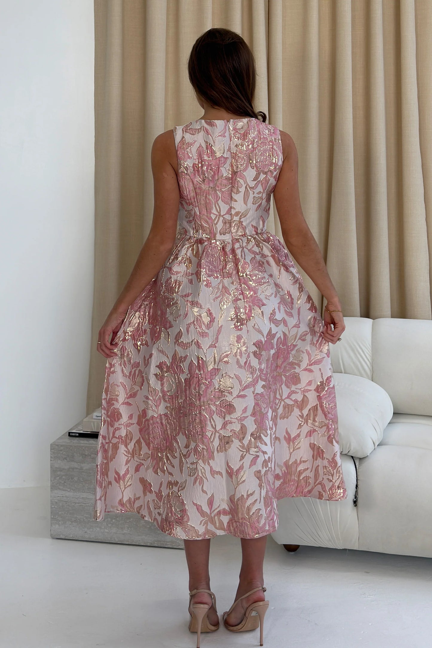 Woman in a floral dress standing in a living room with beige curtains and a white sofa.