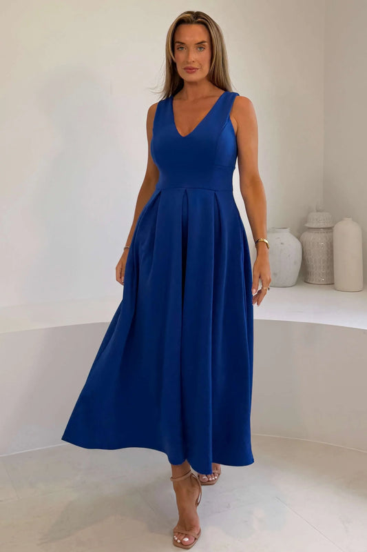 A woman modeling a sleeveless royal blue maxi dress with a V-neckline and a flared skirt, standing in a bright white room.