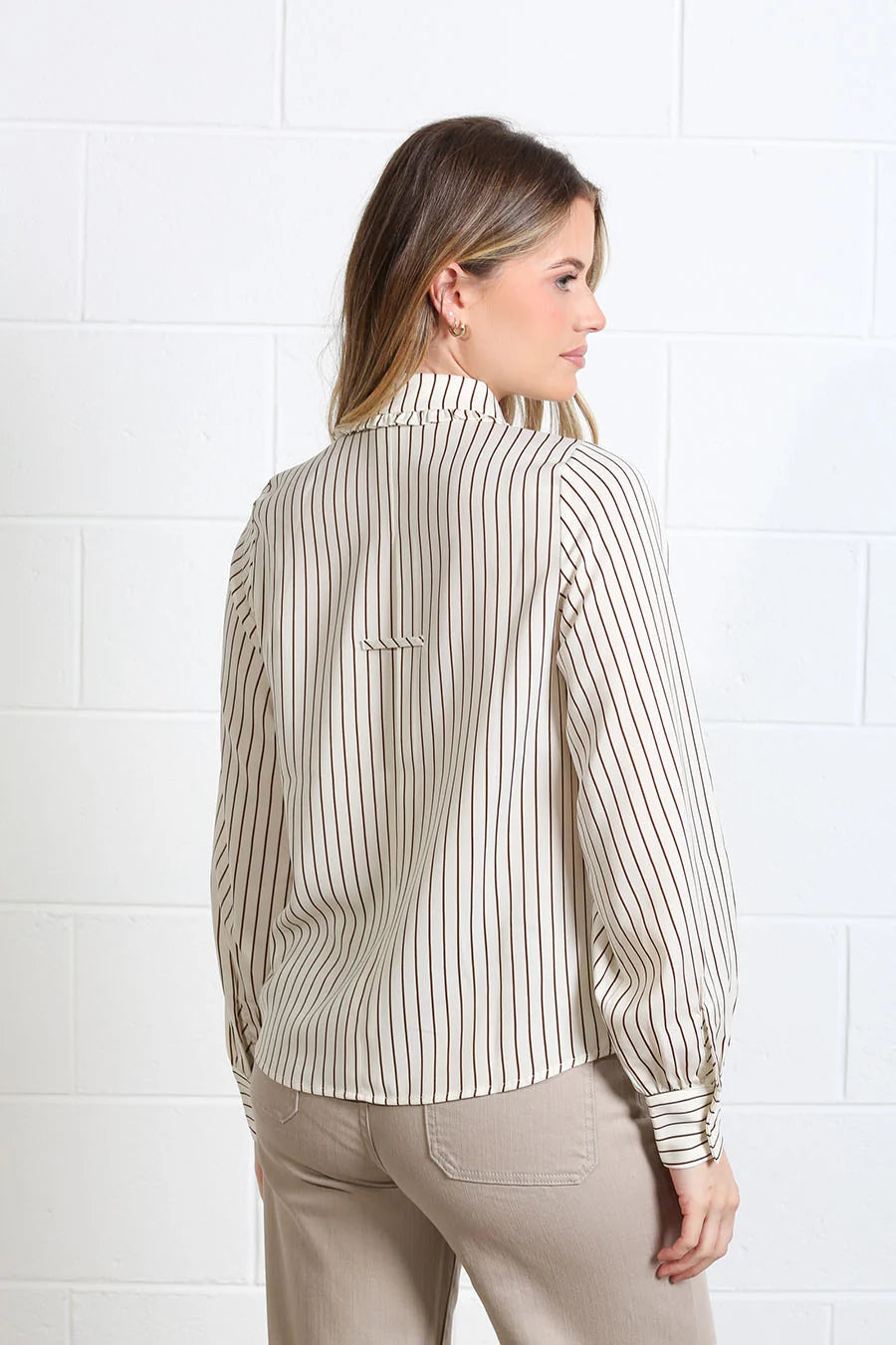 A woman shown from the back wearing a beige and white vertically striped long-sleeve blouse with a high collar, paired with beige pants, standing against a white brick wall.