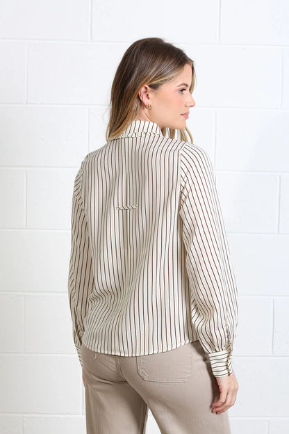 A woman shown from the back wearing a beige and white vertically striped long-sleeve blouse with a high collar, paired with beige pants, standing against a white brick wall.