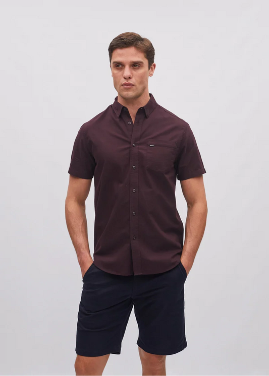 Man wearing a maroon short-sleeve shirt and navy shorts on a light gray background