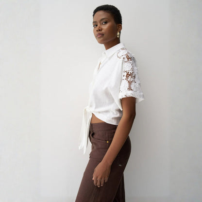 Woman wearing a white blouse with lace sleeves and brown pants against a plain background