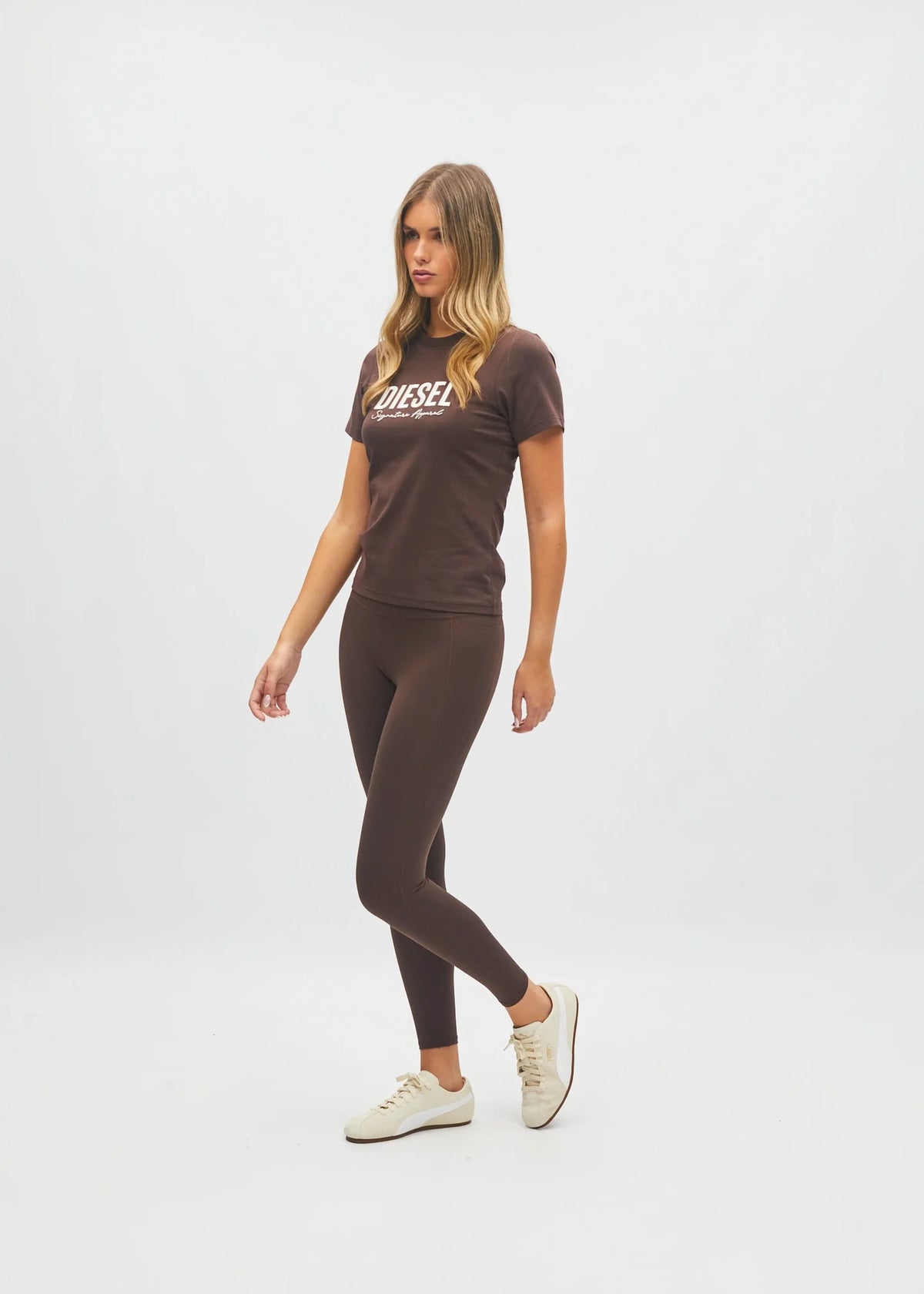 A woman modeling a dark brown Diesel t-shirt and matching dark brown leggings set, paired with off-white sneakers, against a plain light gray studio background.
