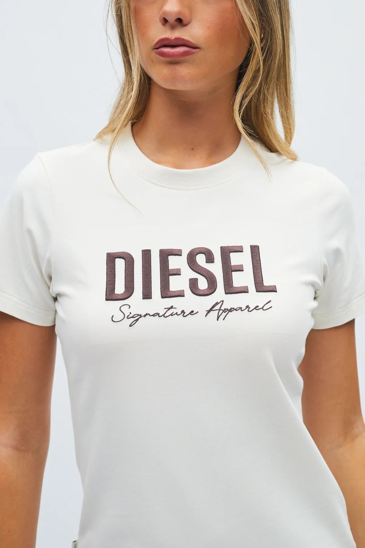 A woman wearing a white short-sleeve crewneck t-shirt with 'DIESEL' and 'Signature Apparel' printed in brown across the chest, shown against a light gray background.