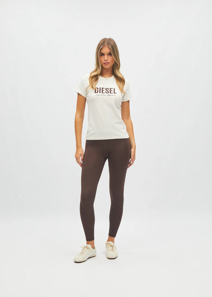 A woman modeling a white short-sleeve t-shirt with 'DIESEL' printed across the chest in brown, paired with dark brown leggings and off-white sneakers, against a plain white studio background.