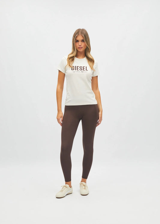 A woman modeling a white short-sleeve t-shirt with 'DIESEL' printed across the chest in brown, paired with dark brown leggings and off-white sneakers, against a plain white studio background.