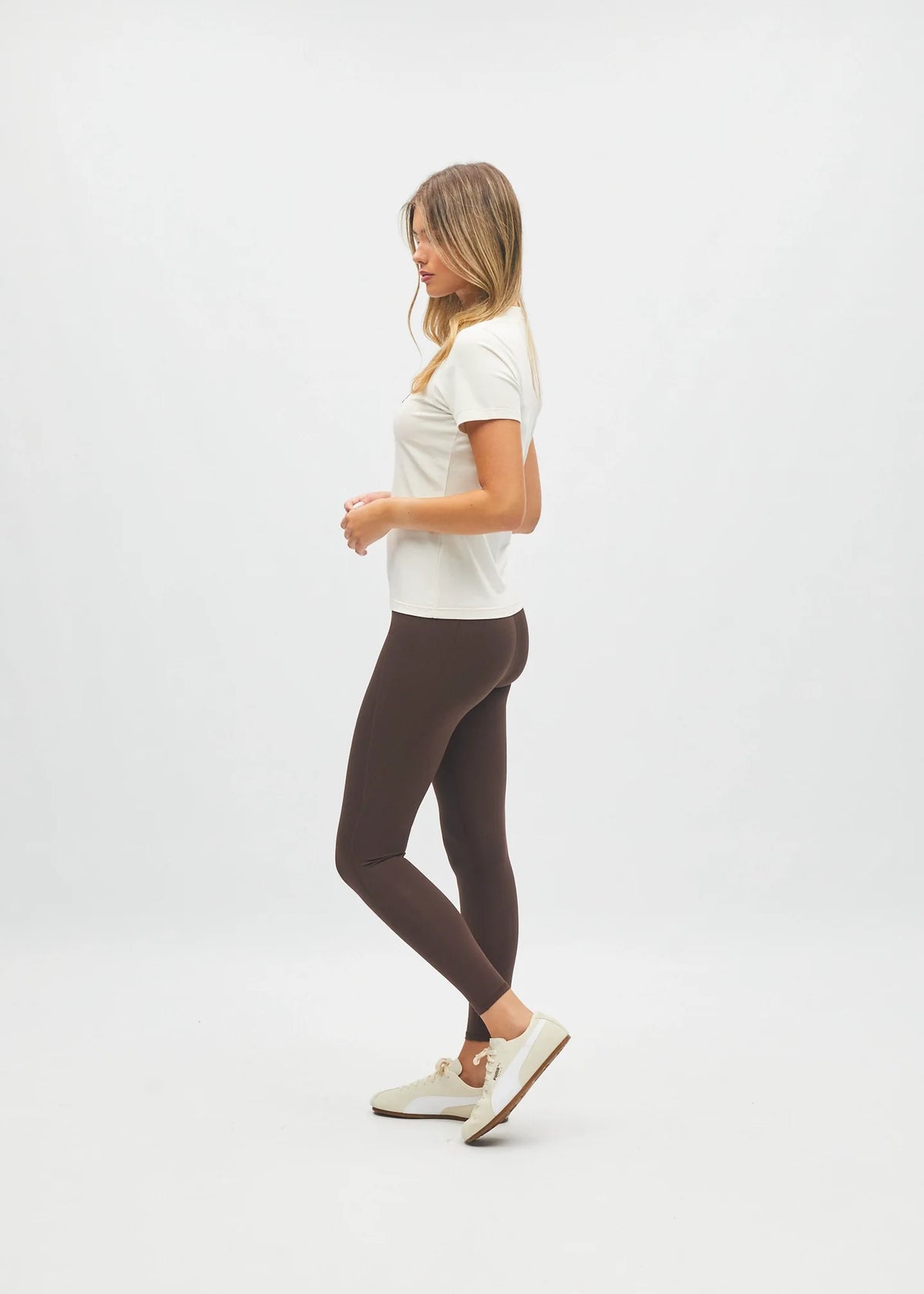 A woman in a white t-shirt and dark brown leggings is shown in profile against a clean white studio background.