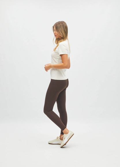 A woman in a white t-shirt and dark brown leggings is shown in profile against a clean white studio background.