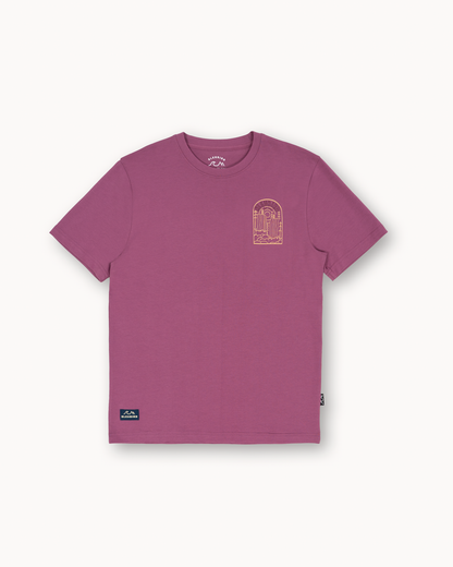 A short-sleeve crewneck t-shirt in a dusty mauve or berry purple color, featuring a small gold graphic on the left chest and a small blue and white logo patch near the bottom left hem, displayed on a clean off-white background.
