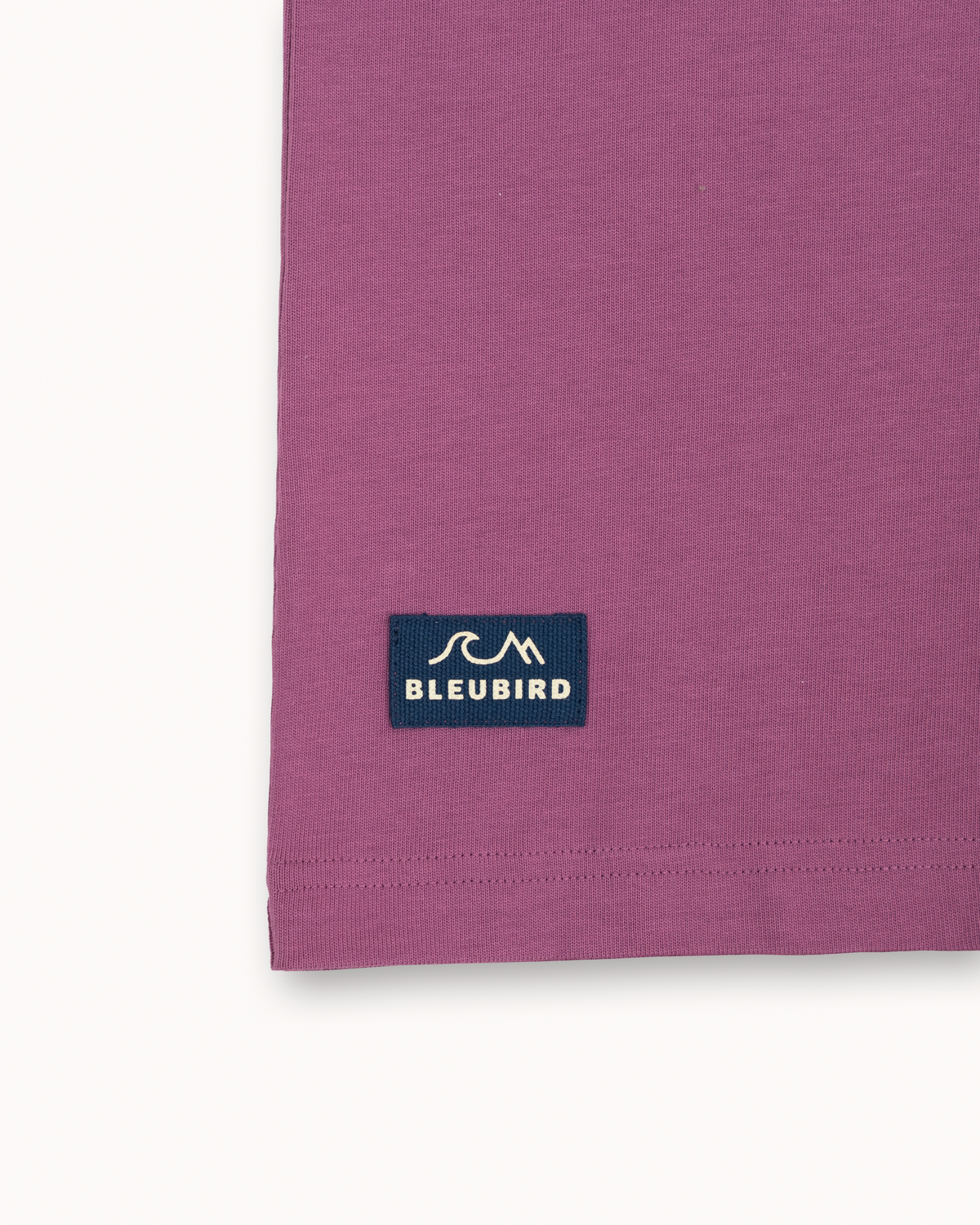 A close-up of a mauve-purple t-shirt corner showing a navy blue 'BLEUBIRD' brand patch with a white logo.