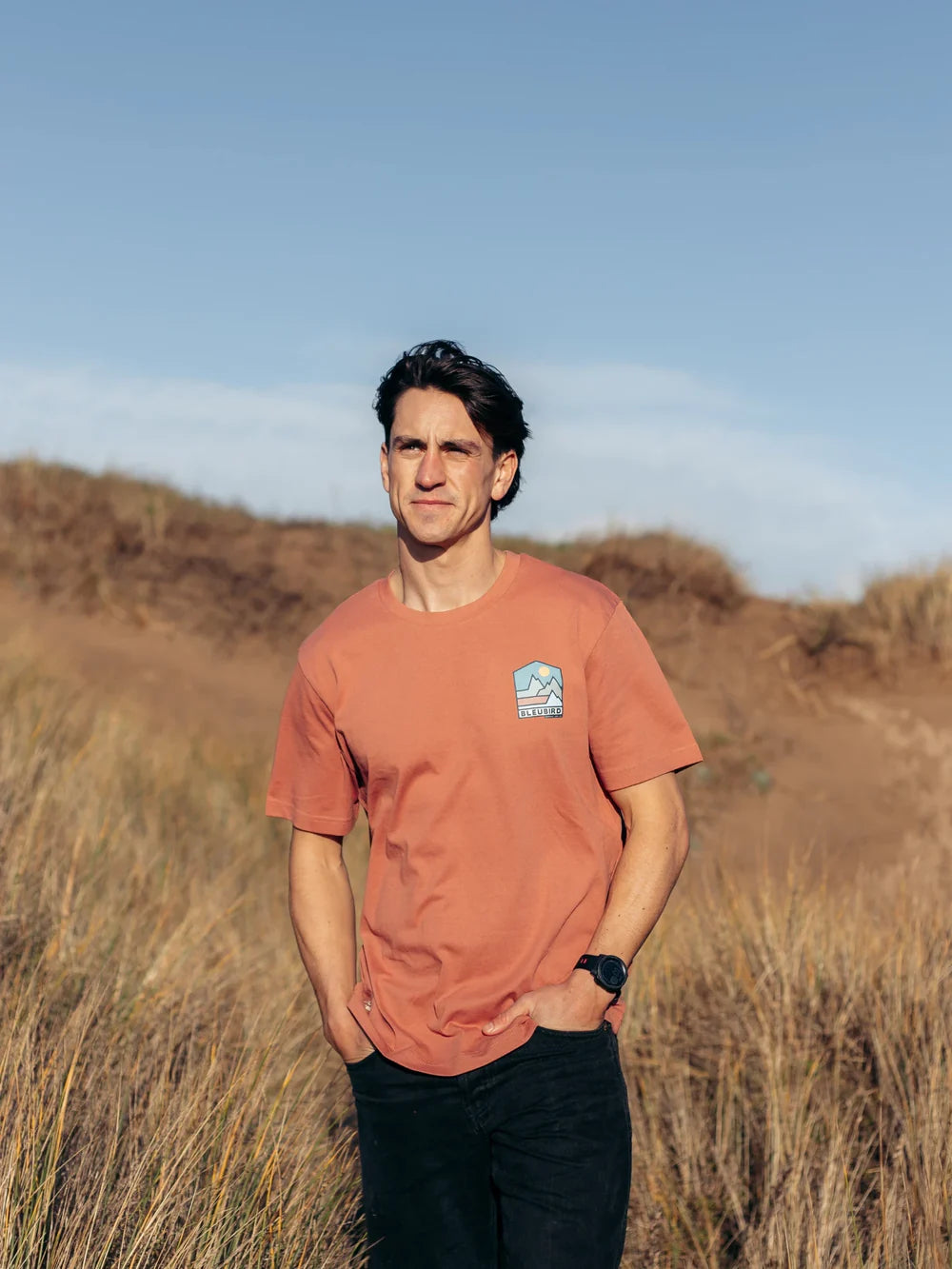 Man wearing an orange t-shirt with a logo standing in a field with a blue sky.
