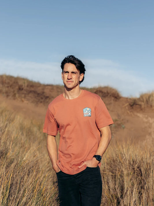 Man wearing an orange t-shirt with a logo standing in a field with a blue sky.