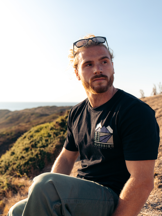 A man sitting outdoors on a hillside wearing a black t-shirt with a white and gray mountain graphic and the brand name 'JANUING' printed on the front, along with gray pants and sunglasses on his head.