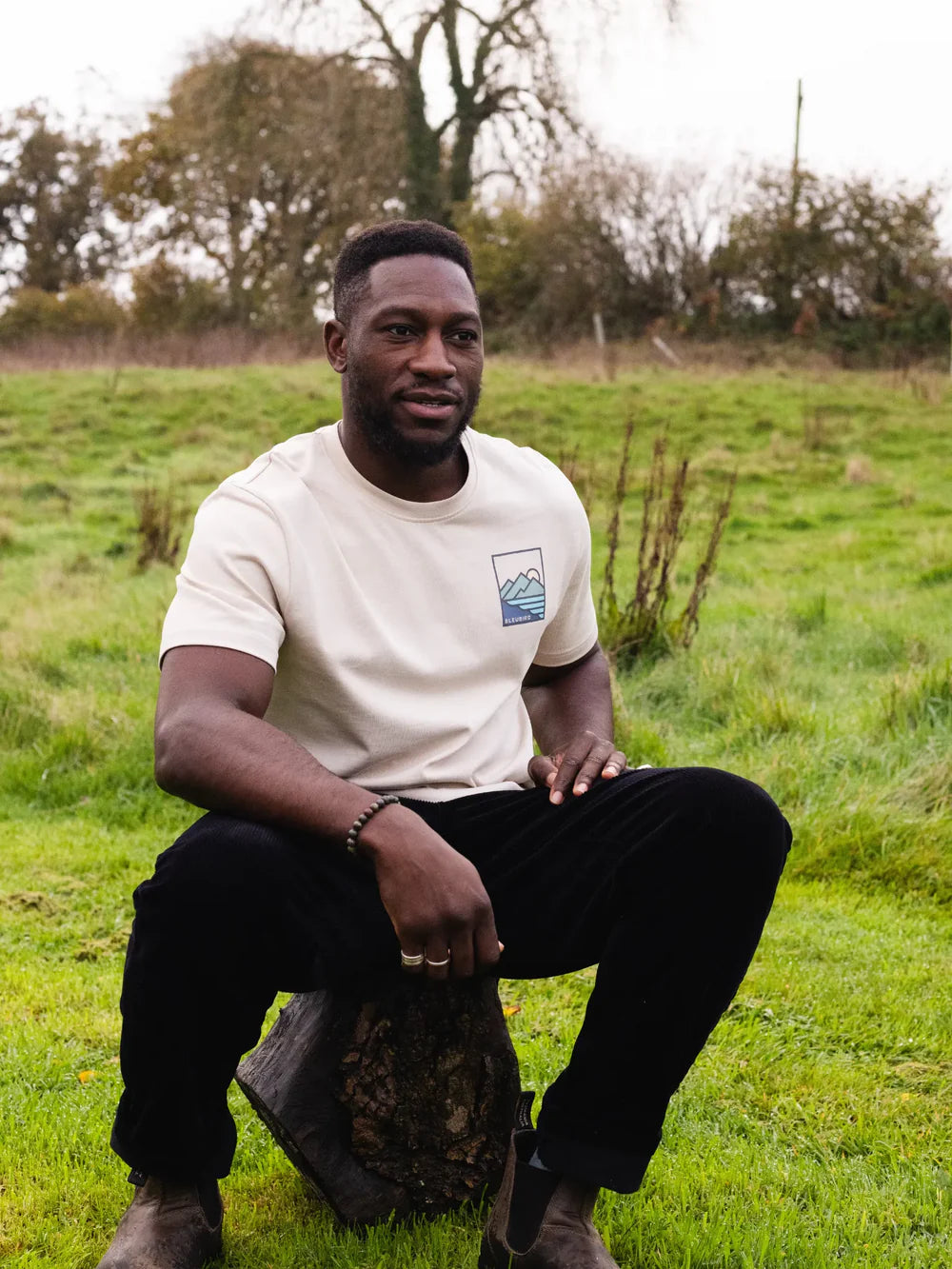 Man sitting on a log in a grassy field wearing a white t-shirt with a logo.