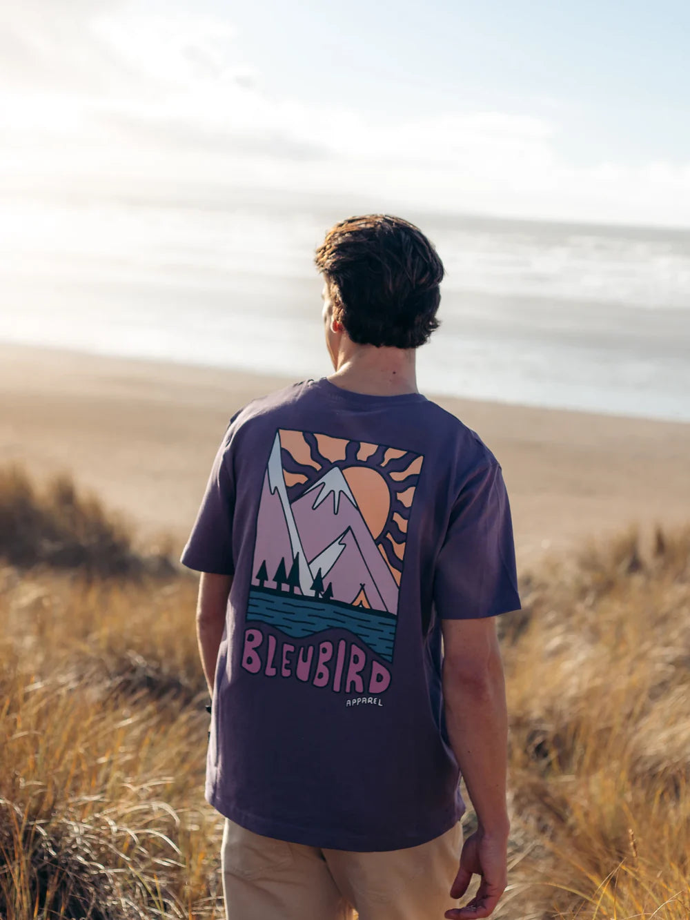 Person wearing a purple t-shirt with a graphic design standing in a field with a sunset in the background