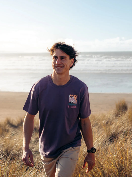 Man walking on a beach wearing a purple t-shirt with a logo.