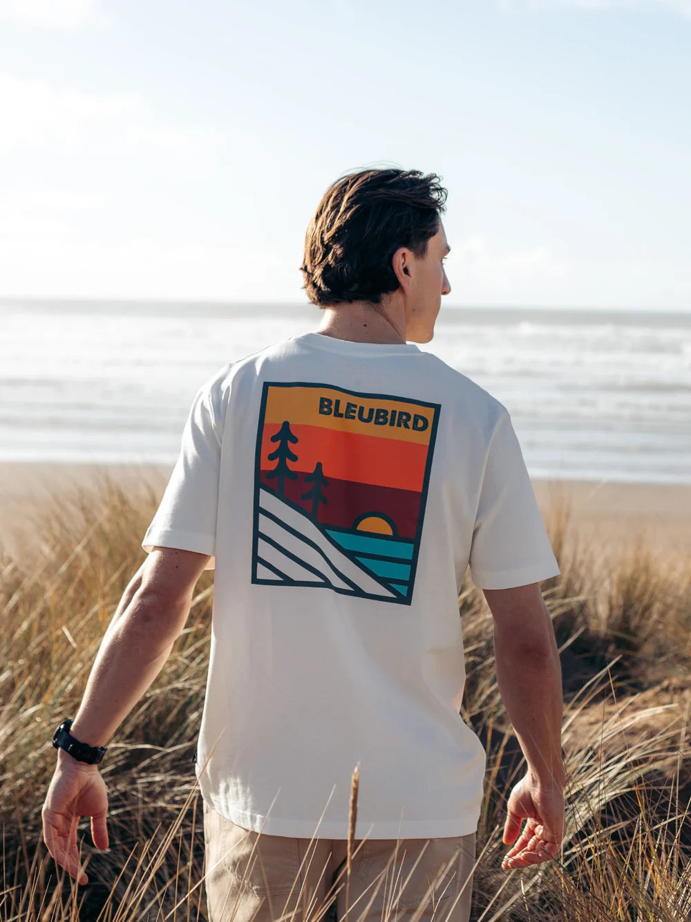 Person wearing a t-shirt with a scenic design and 'BLEUBIRD' branding, standing on a beach.
