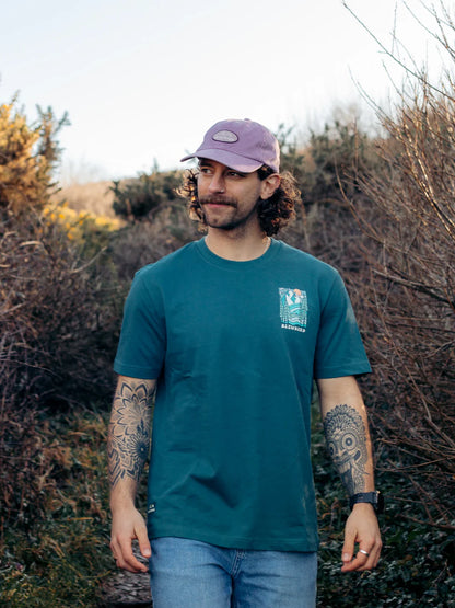 Man wearing a teal t-shirt with a logo, standing outdoors among bushes.