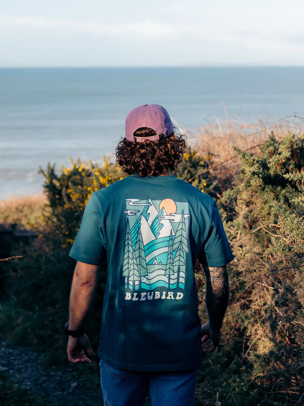 Person wearing a green t-shirt with a logo and text, standing outdoors near a body of water.