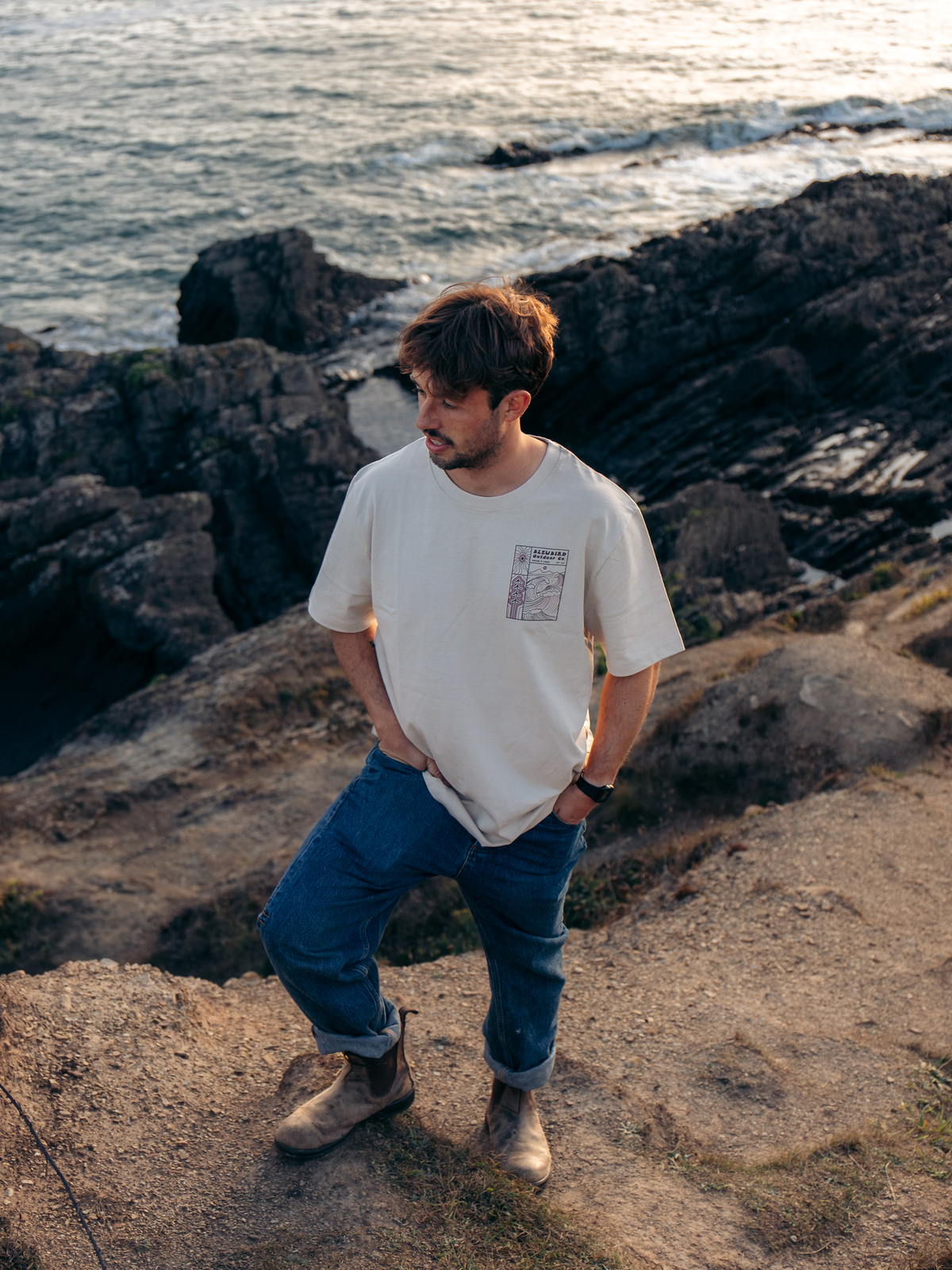 A man stands on a rocky cliffside overlooking the ocean, wearing a cream-colored crewneck t-shirt with a small graphic print on the left chest, blue jeans, and brown boots.