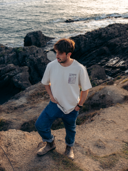 A man stands on a rocky cliffside overlooking the ocean, wearing a cream-colored crewneck t-shirt with a small graphic print on the left chest, blue jeans, and brown boots.
