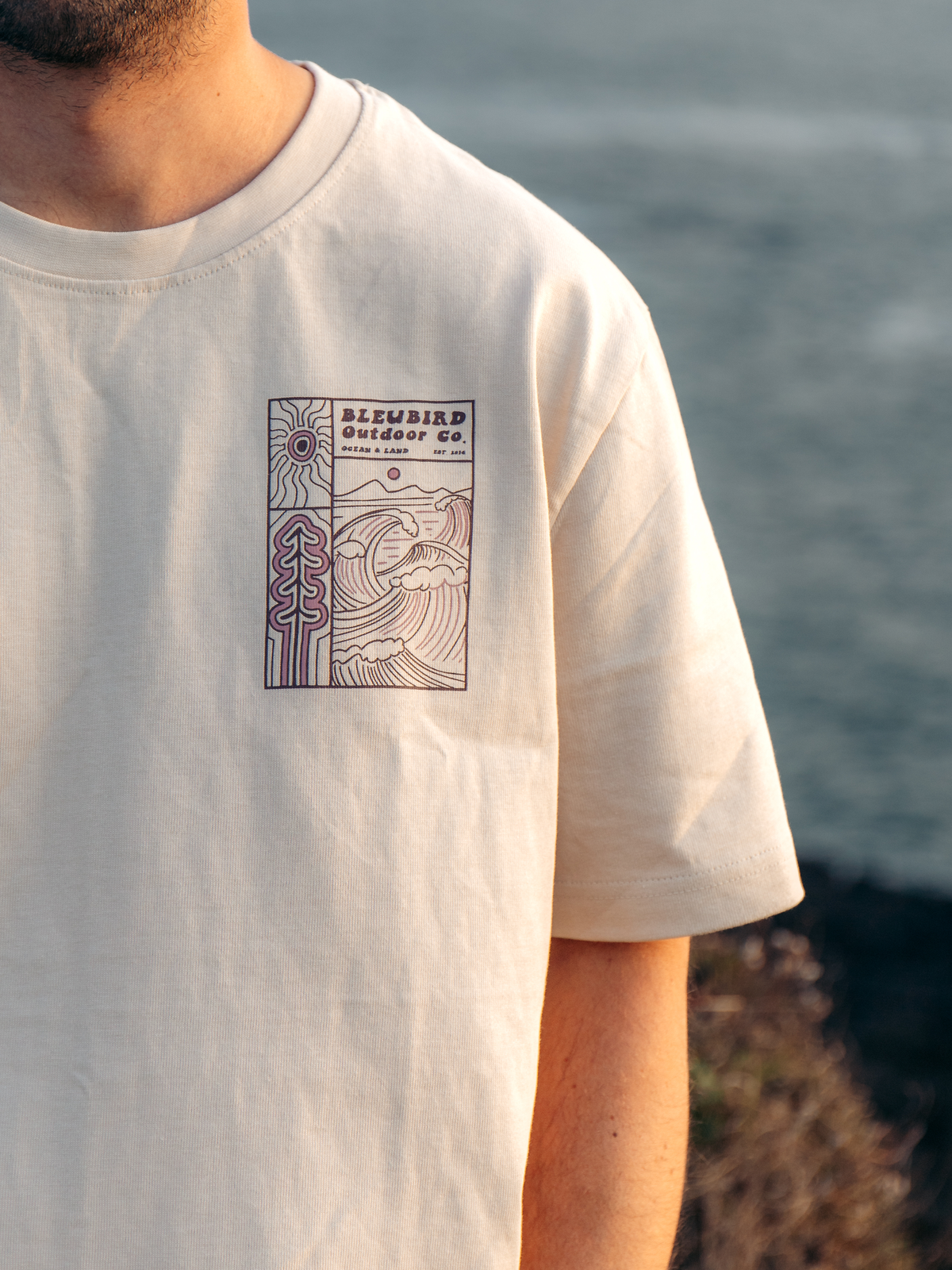 A close-up of a person wearing a cream-colored t-shirt with a purple and brown graphic print on the left chest. The graphic features the text 'BLEYBIRD Outdoor Co' and an illustration of a wave and trees.