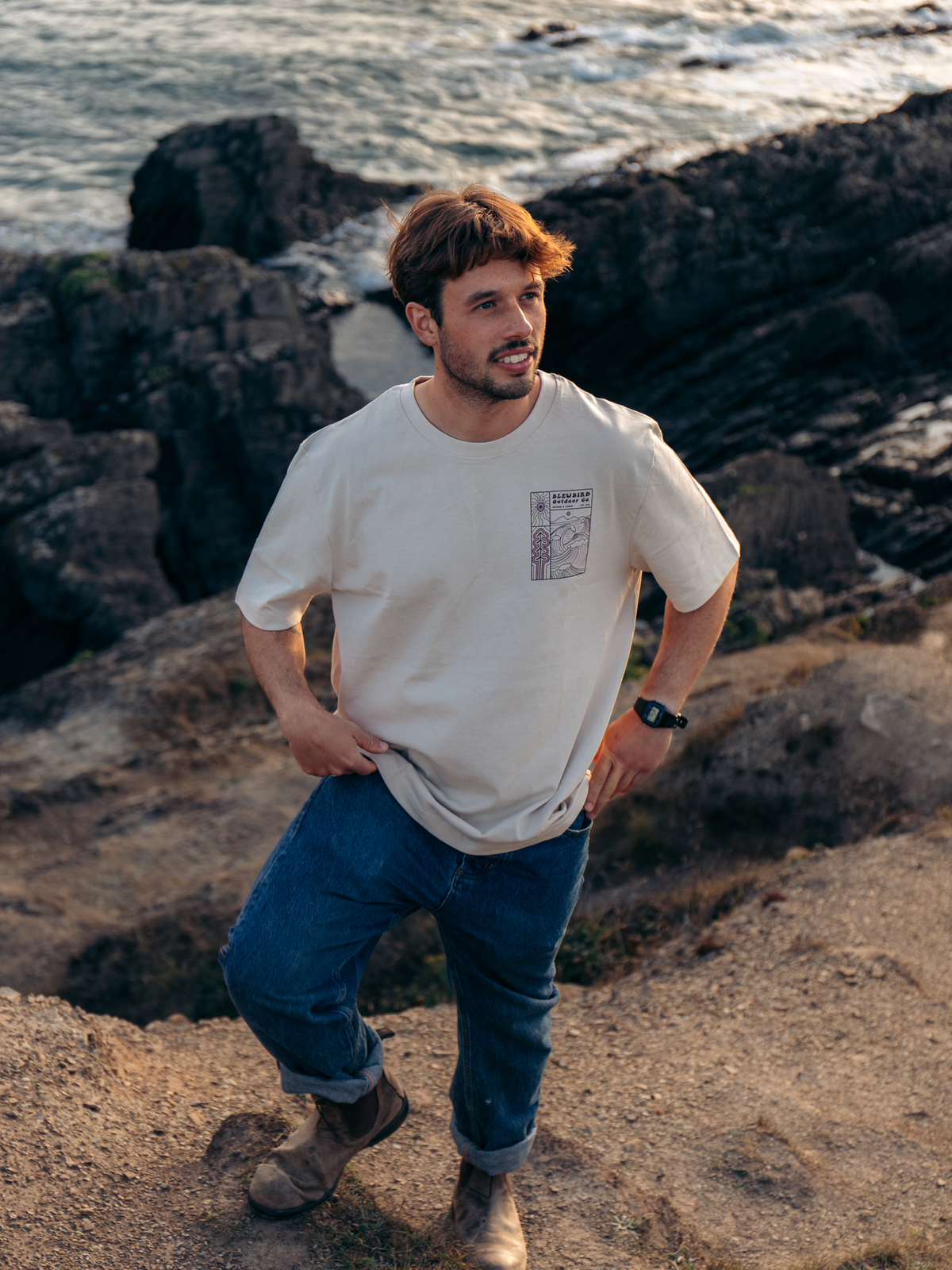 A man wearing a cream-colored graphic t-shirt and blue jeans stands on a rocky cliffside overlooking the ocean at sunset.