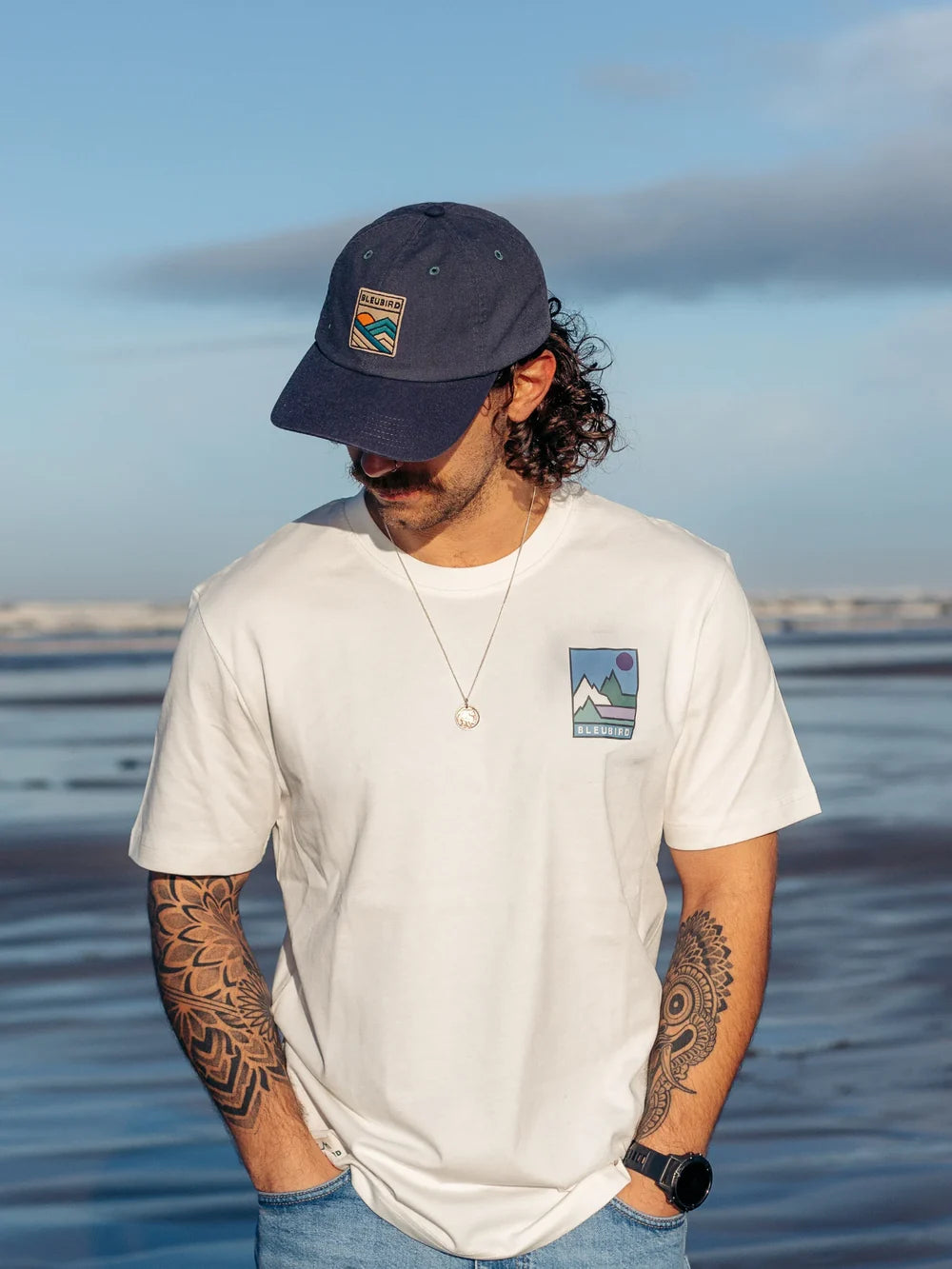 Man wearing a white t-shirt with a logo and a navy cap, standing by a body of water.