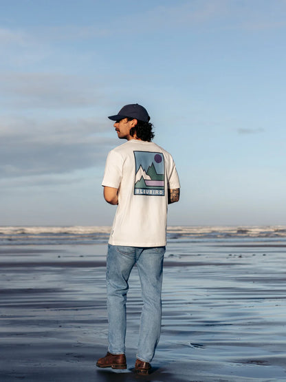 Man standing on a beach wearing a t-shirt with a graphic design, looking out towards the ocean.