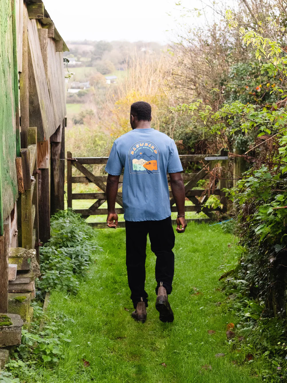 Person walking on a grassy path in a garden wearing a blue t-shirt with a logo.