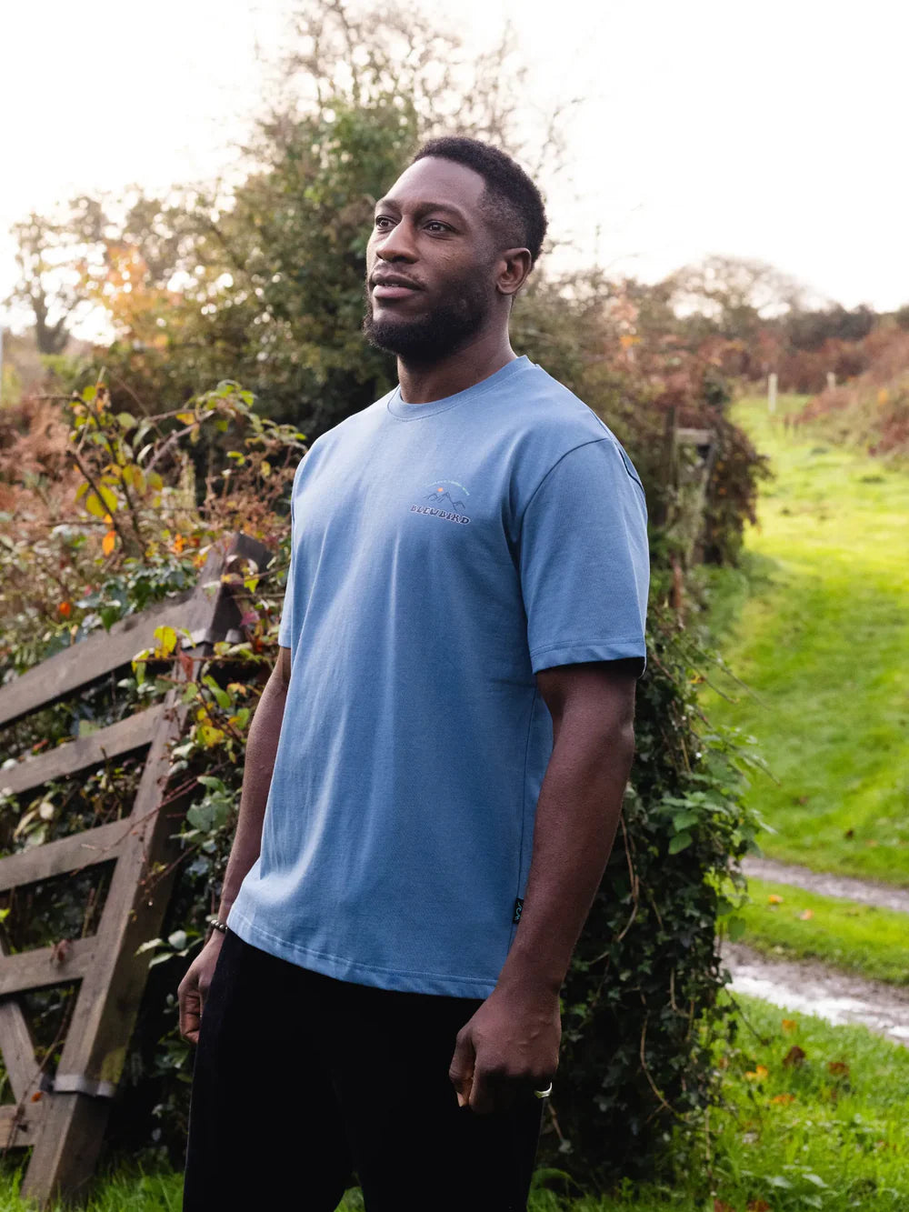 Man wearing a blue t-shirt standing outdoors near a wooden gate and grassy area.
