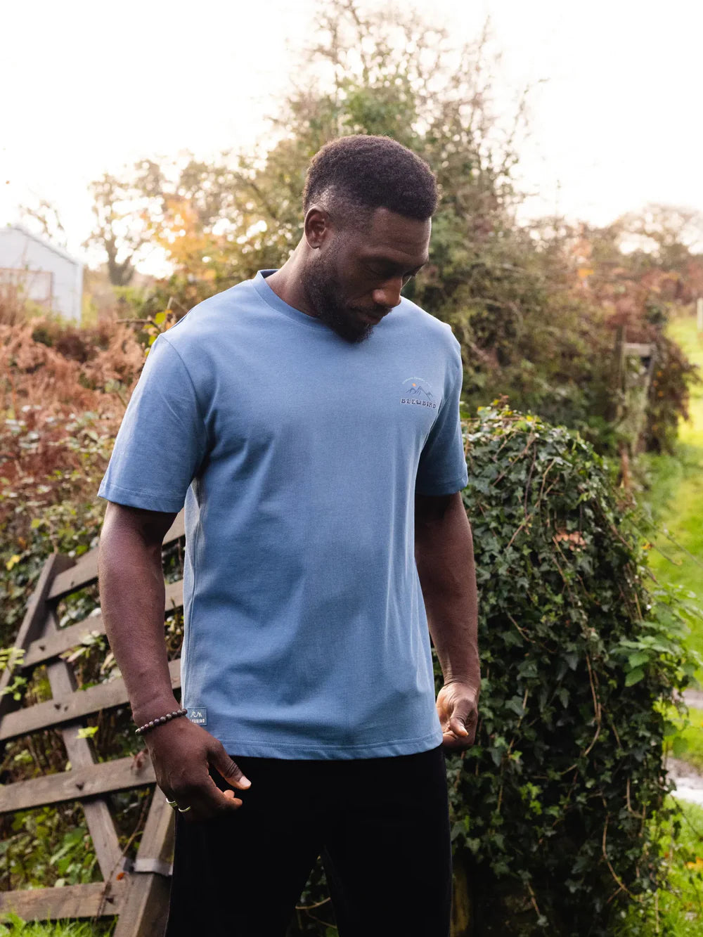 Man wearing a blue t-shirt standing outdoors with greenery in the background