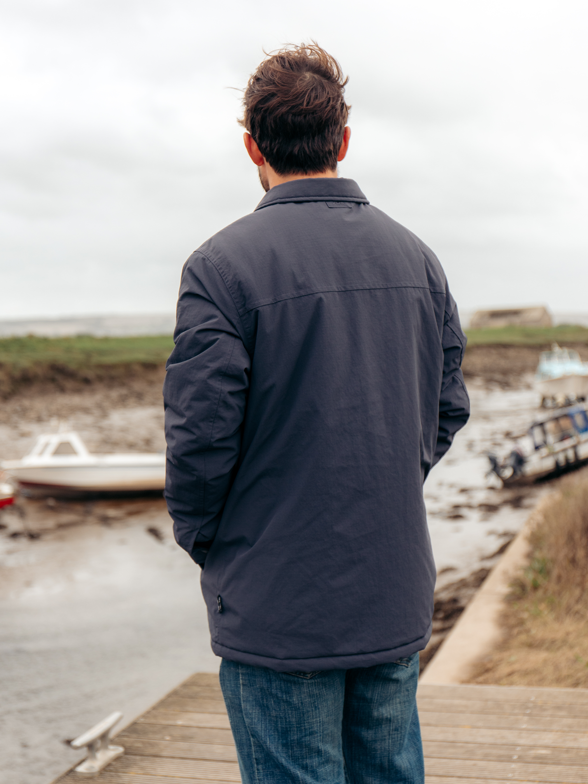 A man viewed from behind wearing a dark navy blue jacket and blue jeans, standing on a wooden dock looking out towards boats on the water under an overcast sky.