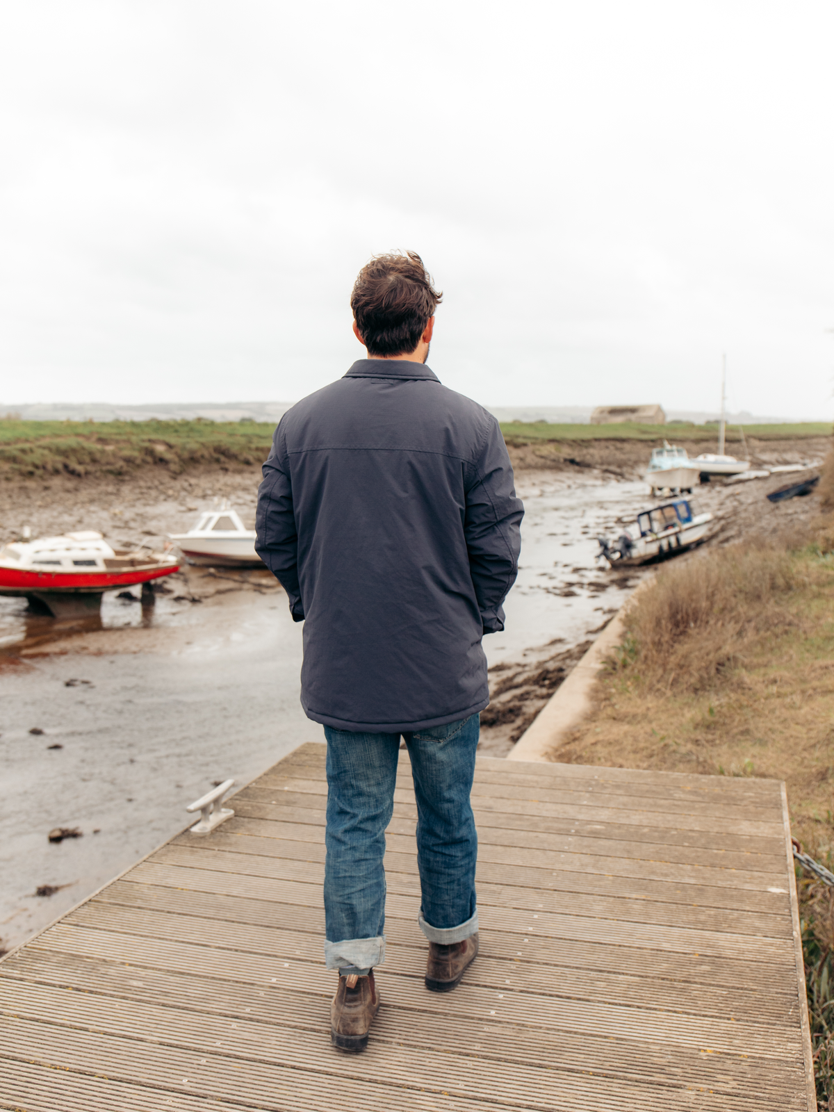 A man viewed from behind stands on a wooden boardwalk overlooking a tidal creek with boats on the mudflats under an overcast sky.