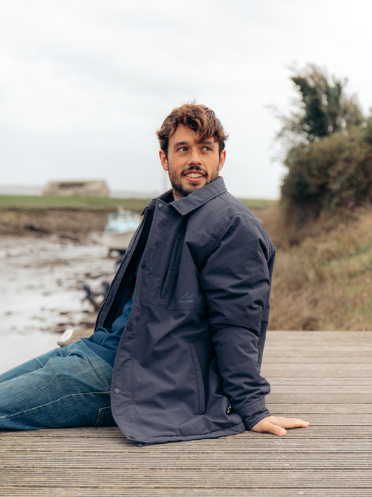 A man wearing a dark navy blue waterproof jacket and blue jeans sits on a wooden boardwalk outdoors with a coastal landscape in the background.
