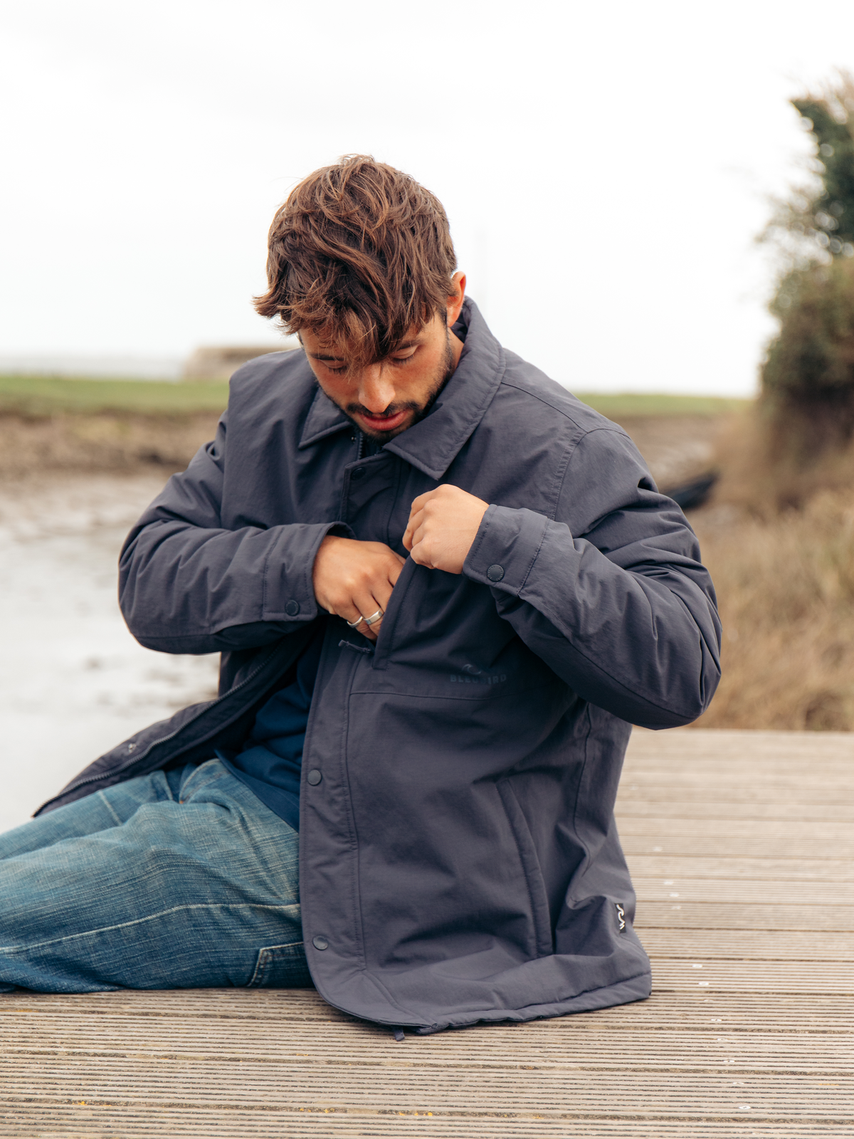 A man wearing a dark slate blue overshirt and blue jeans sits on a wooden boardwalk by the water, looking down while buttoning the overshirt.