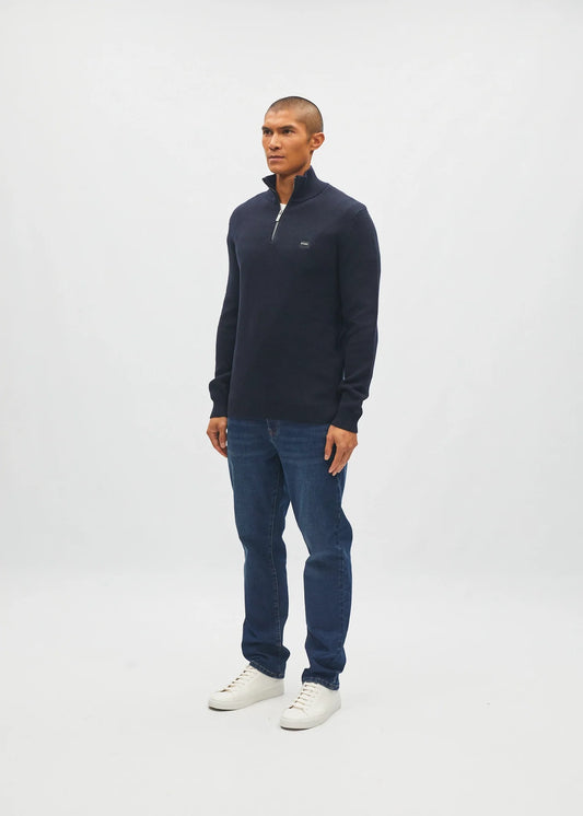 A man modeling a dark navy blue quarter-zip pullover sweater, paired with blue jeans and white sneakers, standing against a light gray studio background.