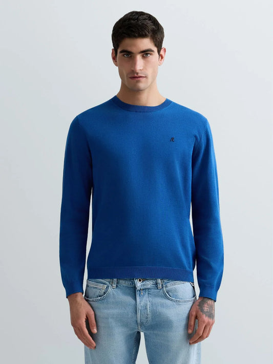 A man wearing a vibrant royal blue crewneck sweater with a small embroidered logo on the left chest, paired with light-wash blue jeans, against a plain light gray studio background.