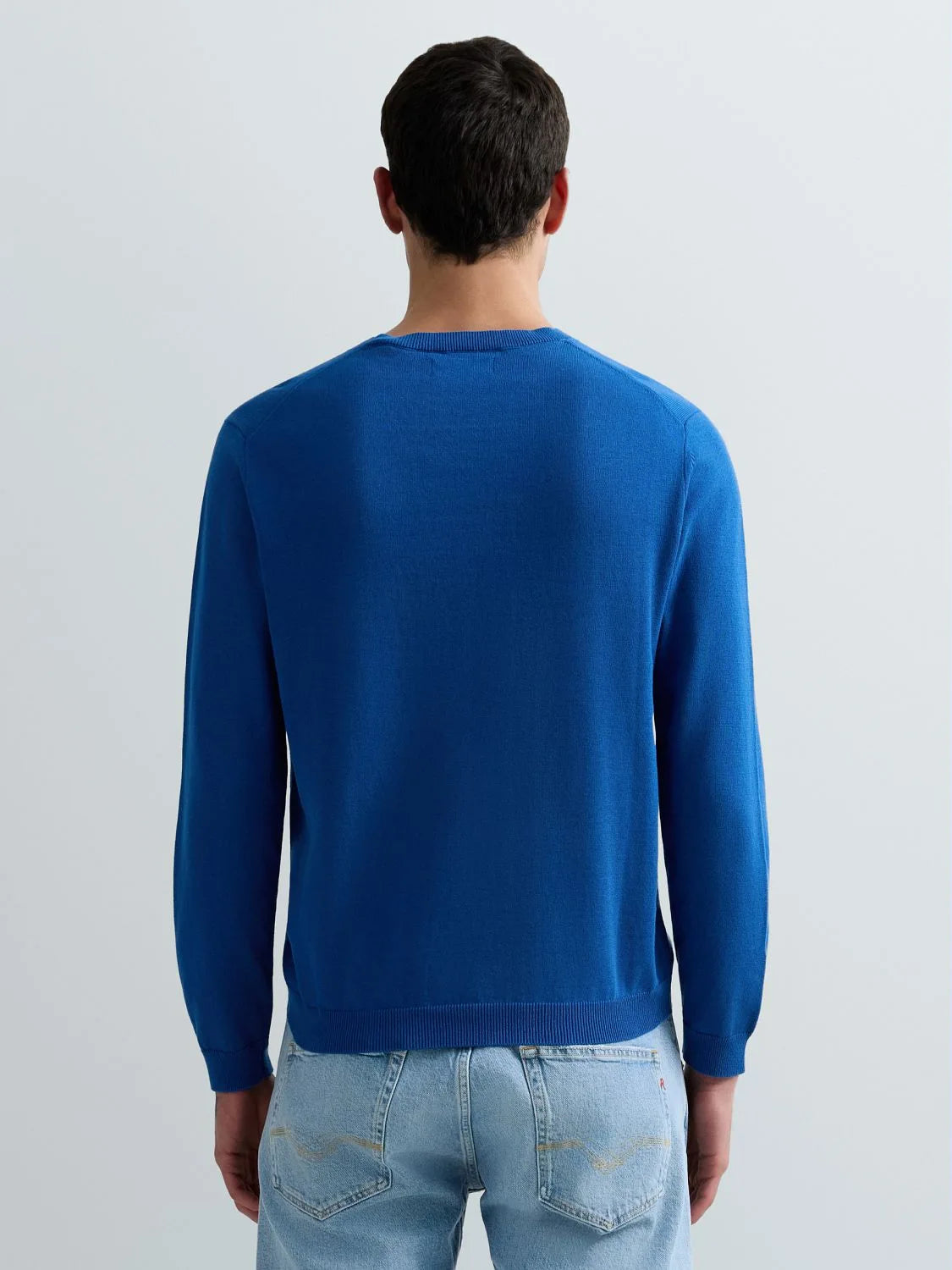 A man viewed from behind wearing a vibrant royal blue long-sleeve knit sweater and light wash blue jeans against a plain light gray studio background.