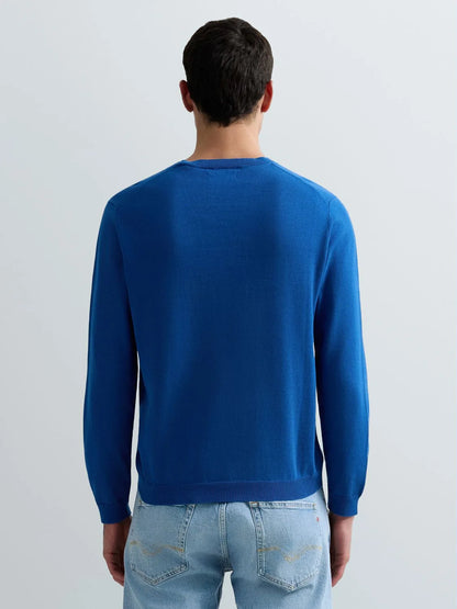 A man viewed from behind wearing a vibrant royal blue long-sleeve knit sweater and light wash blue jeans against a plain light gray studio background.