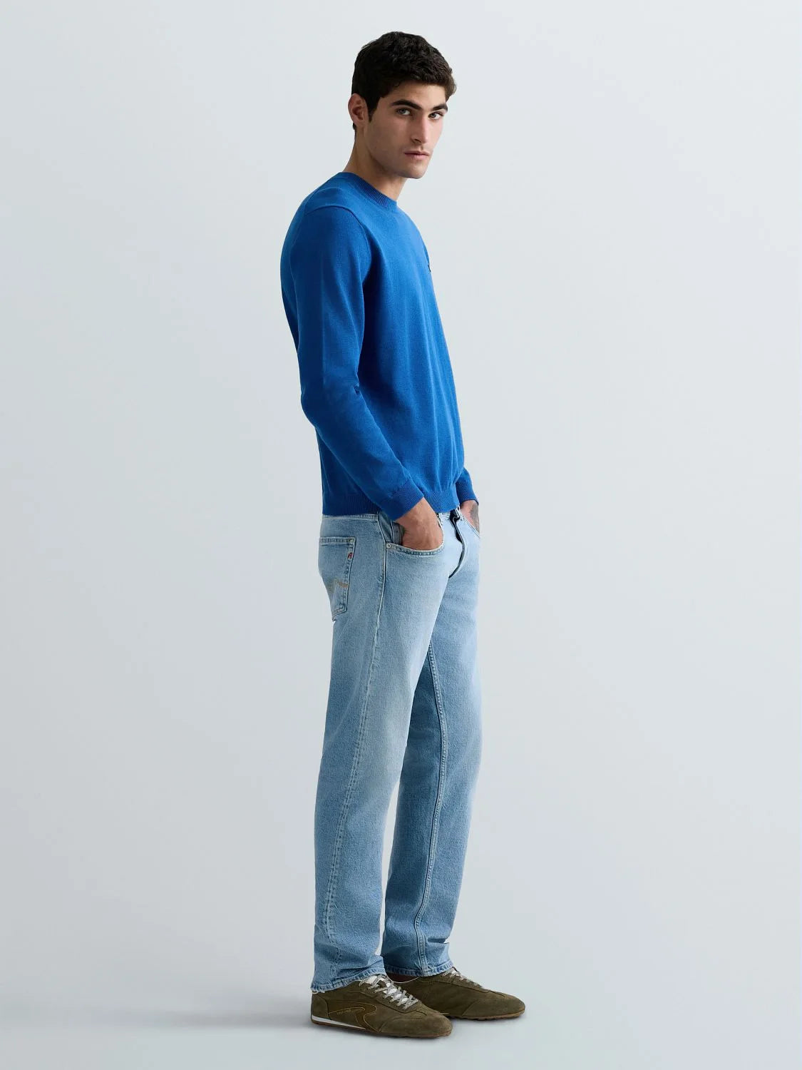 A man wearing a bright blue long-sleeve crewneck sweater, light blue jeans, and brown suede shoes, standing in profile against a light gray studio background.