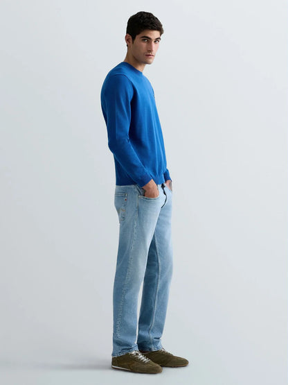 A man wearing a bright blue long-sleeve crewneck sweater, light blue jeans, and brown suede shoes, standing in profile against a light gray studio background.