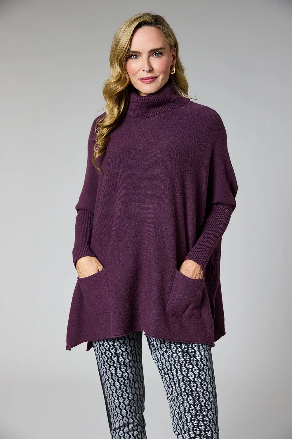 A woman modeling a dark purple turtleneck poncho sweater with front patch pockets, paired with gray patterned leggings, against a solid gray studio background.