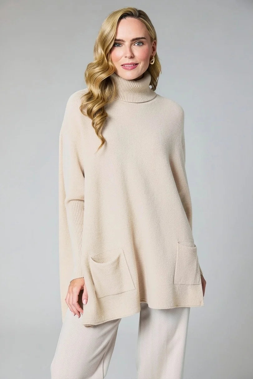 A woman modeling a cream-colored, oversized turtleneck sweater with front patch pockets, paired with matching cream pants, against a solid light gray studio background.