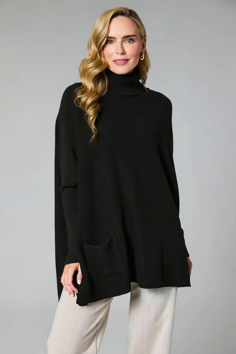 A woman modeling a black oversized turtleneck sweater with front patch pockets, paired with light beige pants, against a solid gray studio background.