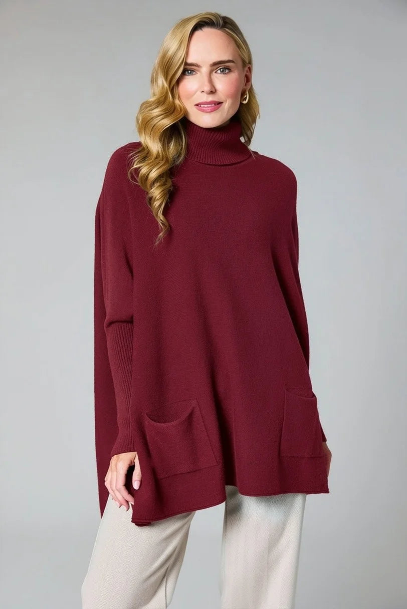 A woman modeling a loose-fitting, long-sleeve turtleneck sweater in deep burgundy with two front patch pockets, paired with light beige pants, against a solid gray studio background.