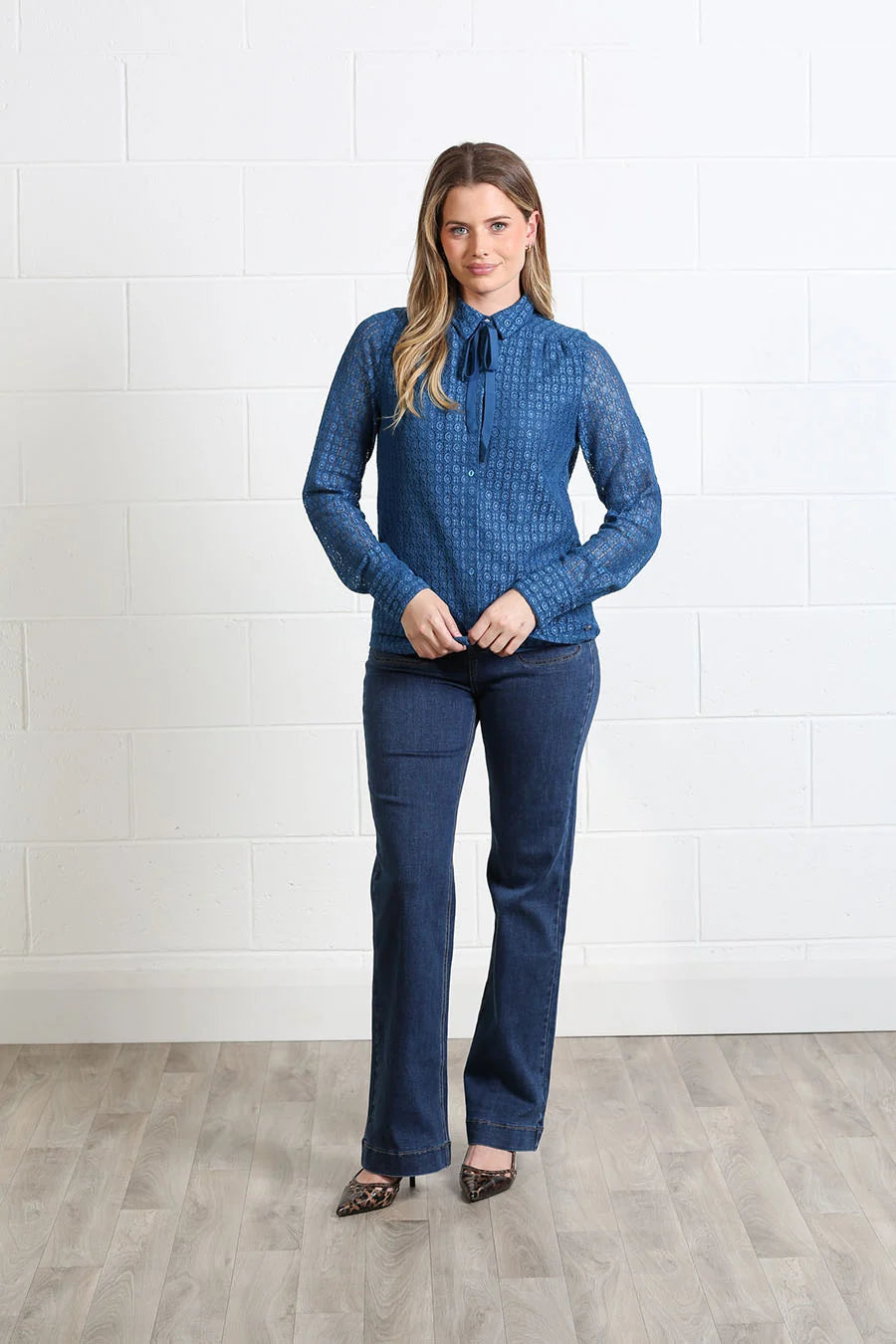 A woman modeling a blue patterned long-sleeve blouse with a tie collar and dark blue flare jeans, standing against a white brick wall and light wood floor.