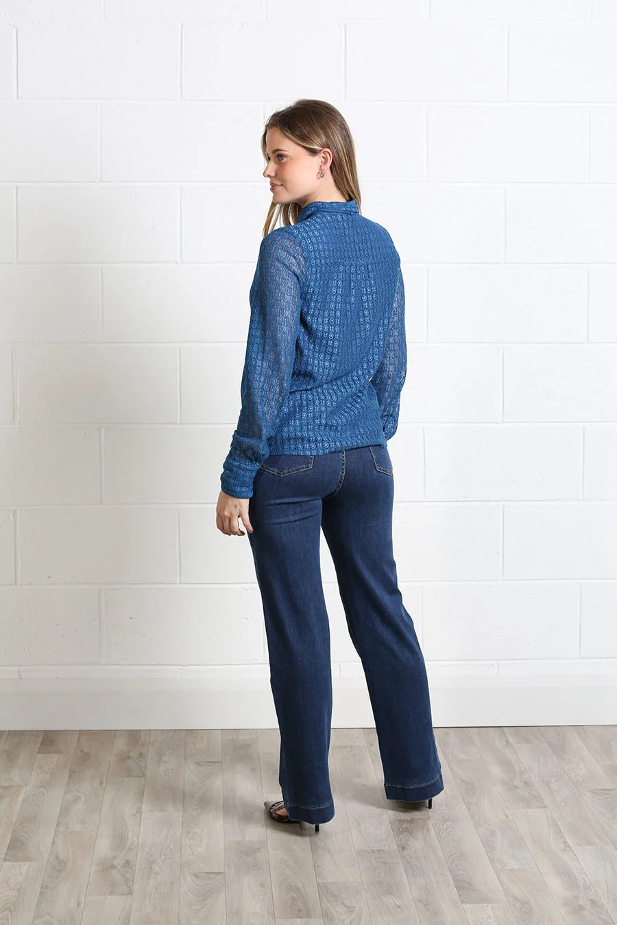 A woman stands in profile wearing a textured blue long-sleeve top and dark blue jeans against a white brick wall and light wood floor.
