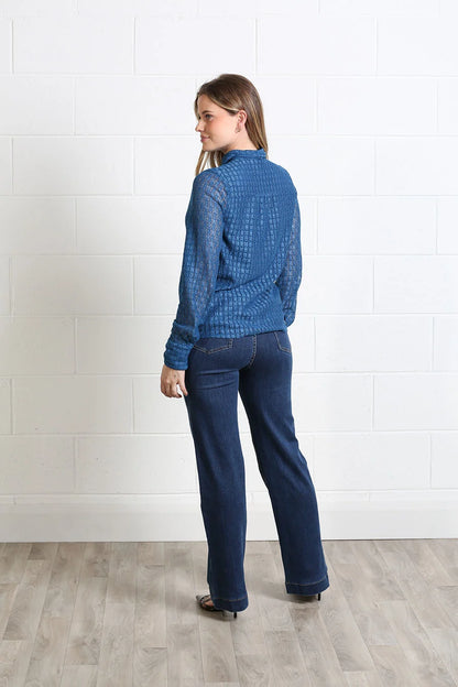 A woman stands in profile wearing a textured blue long-sleeve top and dark blue jeans against a white brick wall and light wood floor.